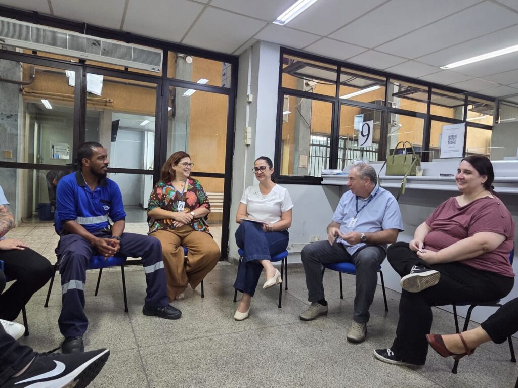 A imagem mostra um grupo de cinco pessoas sentadas em cadeiras azuis dispostas em semi-círculo, em um ambiente interno que parece ser um local público ou escritório. As pessoas estão conversando de forma descontraída e parecem estar confortáveis e engajadas na conversa. Da esquerda para a direita, há um homem vestido com uniforme azul de trabalho, uma mulher com blusa estampada e calça bege, outra mulher com camiseta branca e jeans, um homem com camisa azul clara e calça cinza, e uma mulher com camiseta vinho e calça preta. O ambiente é iluminado com luz branca, o piso é de azulejos claros e há divisórias de vidro ao fundo, mostrando um corredor e uma parede amarela. Ao lado direito da imagem, há uma placa com o número 9 em destaque, provavelmente indicando uma sala ou setor. A cena transmite um clima de reunião informal ou conversa entre colegas.
