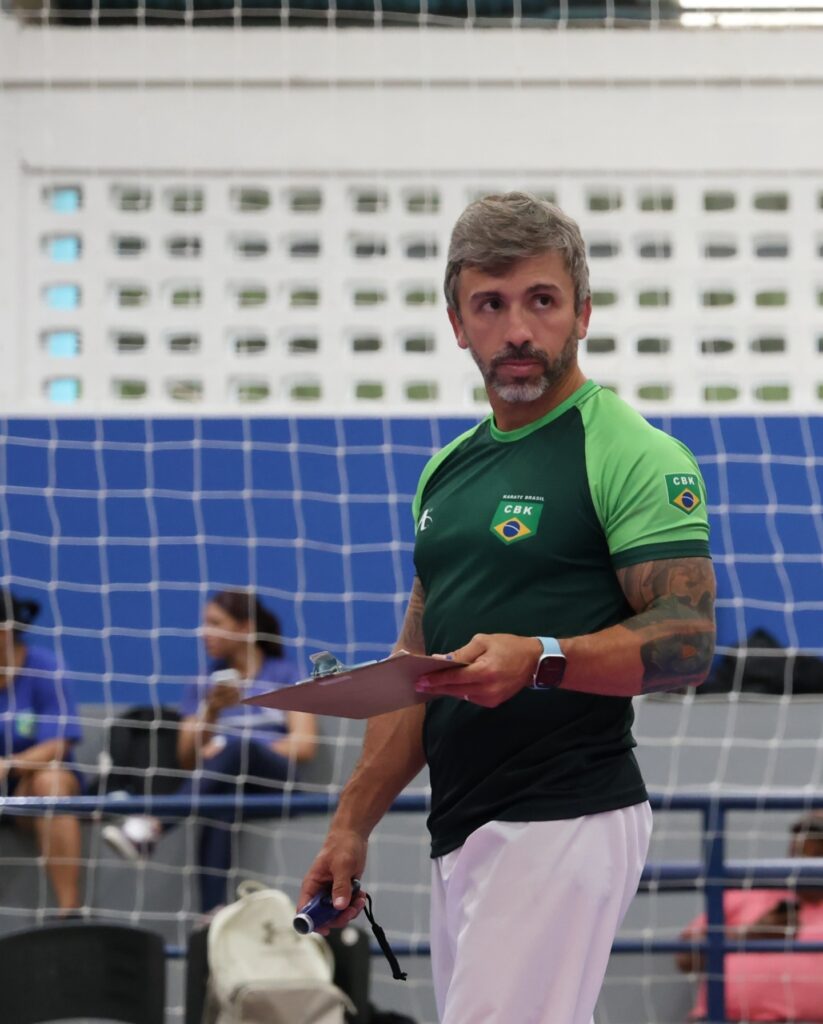 A imagem mostra um homem de meia-idade, provavelmente um treinador ou oficial, em um ambiente esportivo interno, possivelmente uma academia ou ginásio. Ele está usando uma camisa azul clara com o emblema da Confederação Brasileira de Karate (CBK) e a bandeira do Brasil, indicando que ele representa a equipe brasileira de karatê. O homem segura uma prancheta marrom em uma mão e um dispositivo eletrônico azul na outra, que pode ser um cronômetro. Ele tem cabelo e barba grisalhos e vários tatuagens visíveis no braço esquerdo. Ao fundo, desfocado, há outras pessoas sentadas, sugerindo um evento esportivo em andamento. A cena transmite uma atmosfera de concentração e preparação, típica de competições ou treinamentos esportivos.