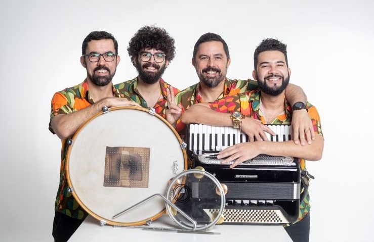 A imagem mostra quatro homens posando juntos atrás de uma mesa branca, cada um segurando instrumentos musicais típicos do forró, um gênero musical brasileiro. Eles estão em um estúdio com fundo branco, o que destaca seus rostos e roupas coloridas. Da esquerda para a direita: o primeiro homem usa óculos e uma camisa laranja com estampa floral; o segundo, com cabelo encaracolado e também de óculos, faz o sinal de paz com a mão; o terceiro, que tem barba e usa um relógio dourado, está com o braço em volta do quarto homem; o quarto homem sorri amplamente. Na frente deles, sobre a mesa, estão os instrumentos: um zabumba (um tipo de tambor grande com a marca "REMO" visível), um triângulo e uma sanfona preta. A imagem transmite uma sensação de alegria, descontração e união, provavelmente de um grupo musical pronto para tocar.