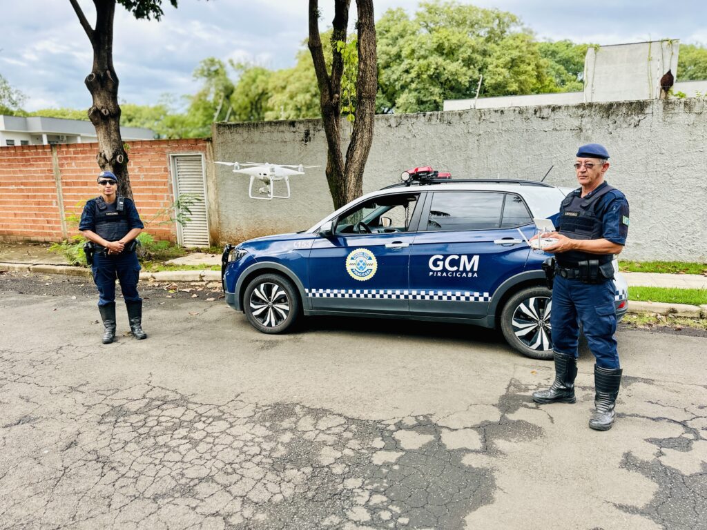 A imagem mostra dois agentes da Guarda Civil Municipal (GCM) de Piracicaba em uma rua asfaltada, que apresenta algumas rachaduras. À direita, um agente masculino mais velho está em destaque, vestindo uniforme azul escuro, boina azul, colete tático e óculos. Ele segura um controle remoto de drone branco. À esquerda, uma agente feminina mais jovem está próxima a uma árvore, também vestindo uniforme e colete tático, além de óculos escuros, com as mãos cruzadas à frente. Entre eles, há uma viatura da GCM, um Volkswagen T-Cross com cores azul e branca, e inscrições "GCM PIRACICABA" e o brasão da prefeitura na porta. Acima da viatura, um drone branco de quatro hélices está voando. Ao fundo, há um muro de concreto antigo, uma parede de tijolos com uma porta branca, além de árvores verdes. O cenário é ao ar livre, durante o dia, com iluminação natural. A imagem transmite a ideia de modernização tecnológica da guarda municipal, com o uso de drones para vigilância.