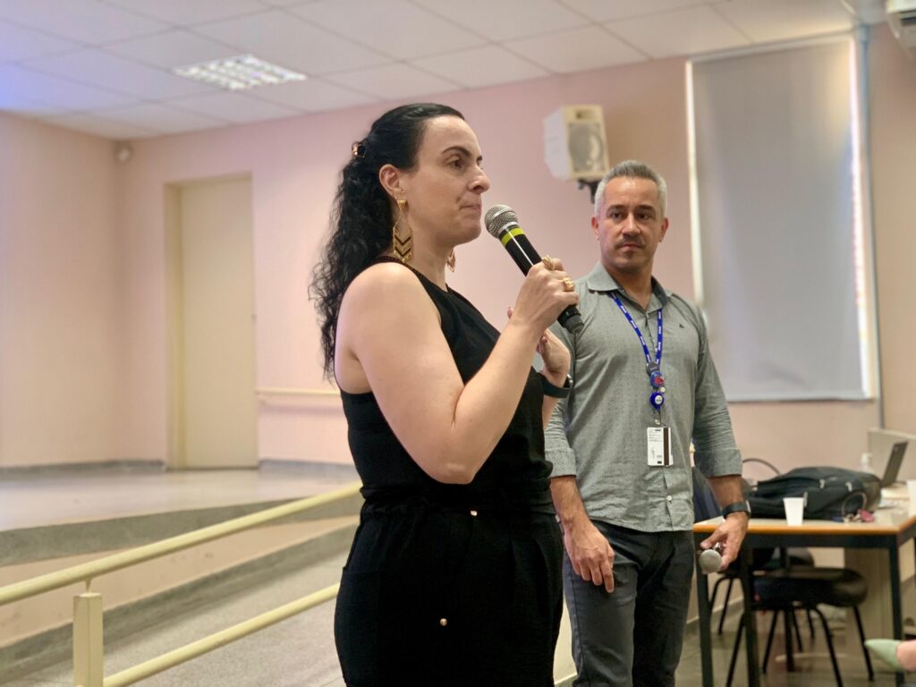 A imagem mostra duas pessoas em um ambiente que parece ser uma sala de aula ou espaço de seminário. A mulher em primeiro plano está falando ao microfone, vestindo uma roupa preta sem mangas e brincos dourados, transmitindo a ideia de estar conduzindo uma apresentação. Ao lado dela, um homem segura outro microfone, usando uma camisa cinza e um crachá pendurado em um cordão, sugerindo que também participa da condução do evento. Atrás deles há uma mesa com um laptop, uma mochila e um copo descartável, reforçando o contexto de trabalho ou estudo. O espaço tem paredes claras, uma janela com persiana fechada e um alto-falante fixado na parede, compondo o cenário de uma atividade educacional ou profissional.