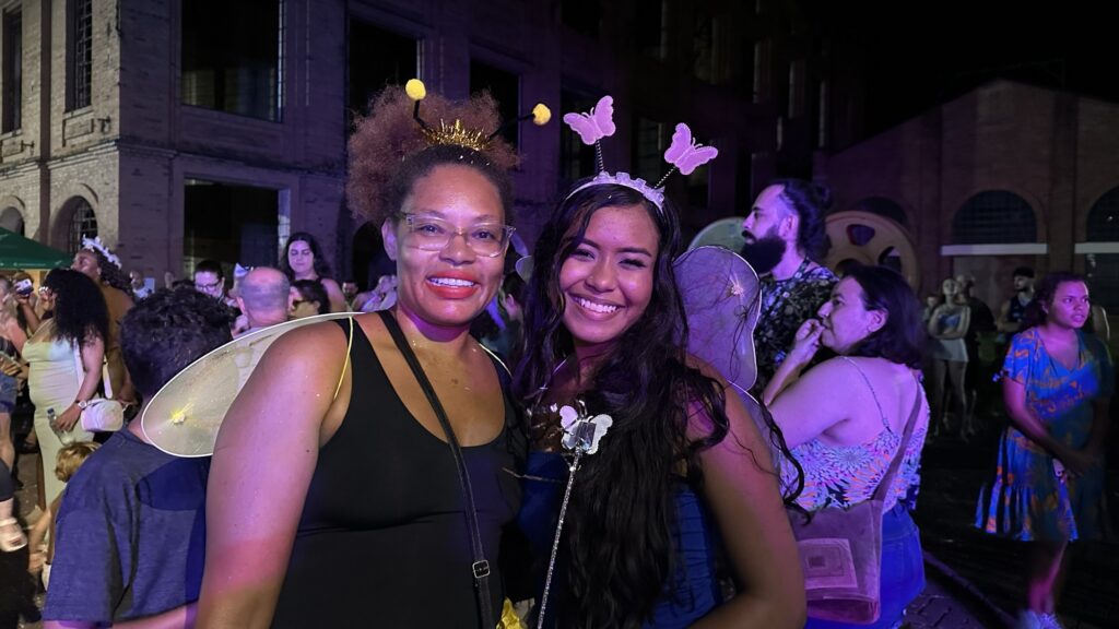 A imagem mostra duas mulheres posando para uma foto durante um evento ao ar livre à noite. Ambas estão usando acessórios de fantasia: a mulher à esquerda está com uma tiara com antenas de abelha amarelas e pretas e asas brancas transparentes; ela usa óculos e uma regata preta. A mulher à direita usa uma tiara com pequenas borboletas rosas em molas e asas combinando, além de uma blusa azul. Elas estão sorrindo e parecem se divertir. Ao fundo, há outras pessoas, algumas delas também parecem estar fantasiadas, e um prédio de tijolos grandes, indicando que o evento ocorre em um espaço aberto, possivelmente um festival ou festa temática. A iluminação é predominantemente roxa, criando um clima festivo e alegre.