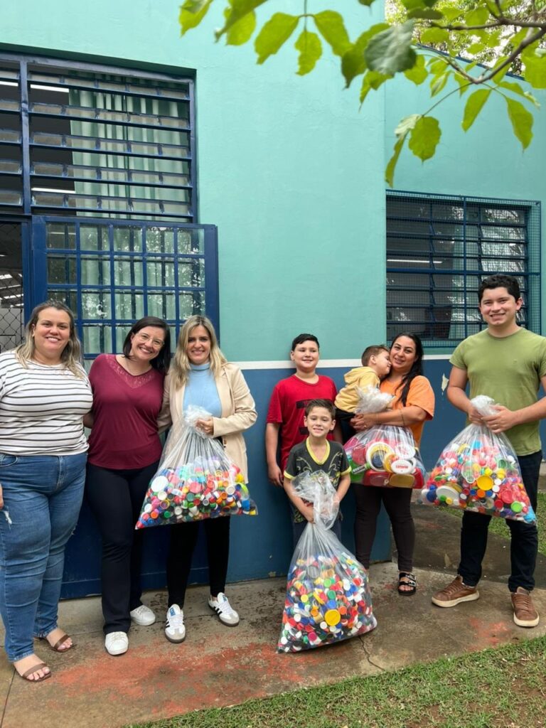 A imagem mostra um grupo de oito pessoas, incluindo adultos e crianças, posando em frente a um prédio azul claro com detalhes em azul escuro. Todos estão segurando grandes sacos transparentes cheios de tampas e recipientes plásticos coloridos. O grupo está alinhado, sorrindo para a foto, transmitindo uma atmosfera de colaboração e alegria. As pessoas parecem estar envolvidas em uma atividade de coleta ou reciclagem de materiais plásticos, sugerindo um esforço comunitário ou ambiental. Ao fundo, o prédio tem janelas com grades e uma faixa branca horizontal, e o ambiente é ao ar livre, com algumas folhas verdes visíveis no canto superior direito da imagem.