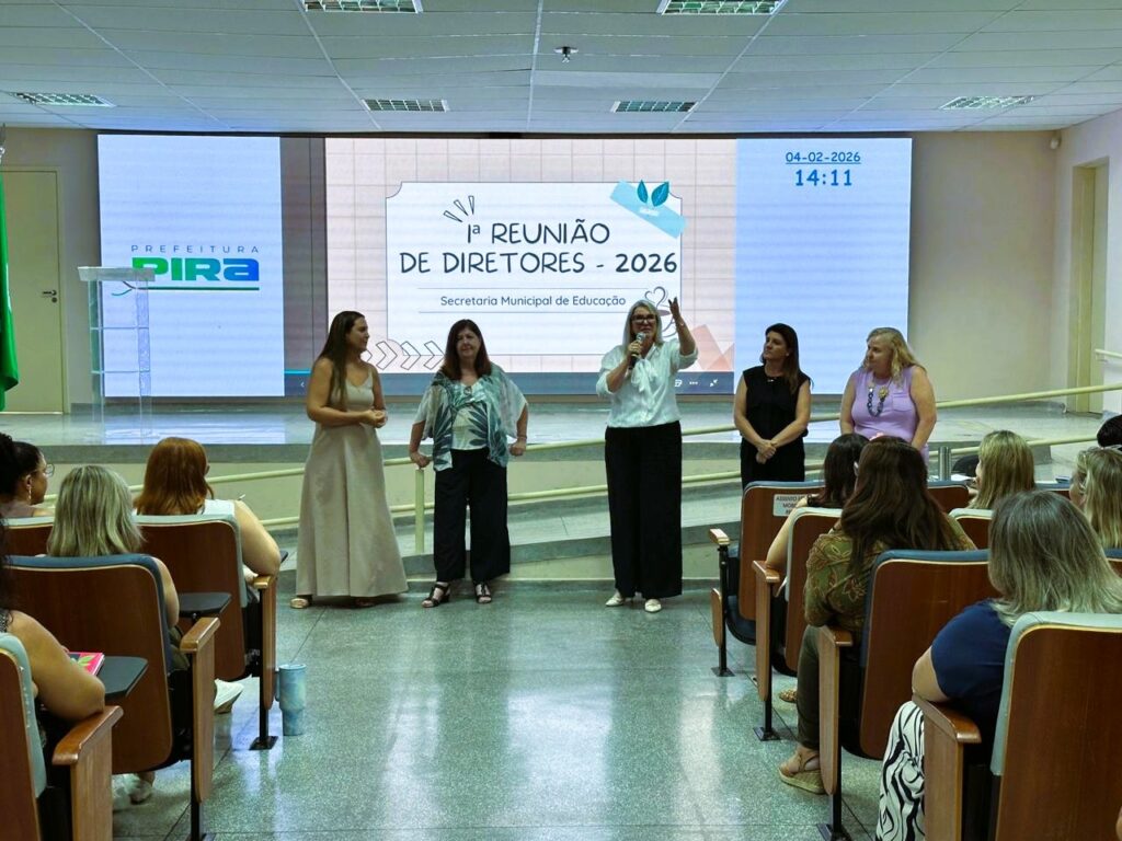 A imagem mostra uma reunião formal realizada em um auditório. No palco, cinco mulheres estão alinhadas, todas com roupas formais, demonstrando um ambiente profissional. Uma delas está falando ao microfone, com a mão direita levantada, provavelmente enfatizando um ponto importante. Atrás delas, há uma grande tela digital que exibe informações em português, incluindo a data "04-02-2026", a hora "14:11" e o título do evento "1ª REUNIÃO DE DIRETORES - 2026", organizada pela "Secretaria Municipal de Educação" da "Prefeitura Pira". Na parte inferior da imagem, é possível ver parte do público, sentado em cadeiras, assistindo à apresentação. O ambiente é bem iluminado e o cenário é típico de uma conferência ou reunião oficial.