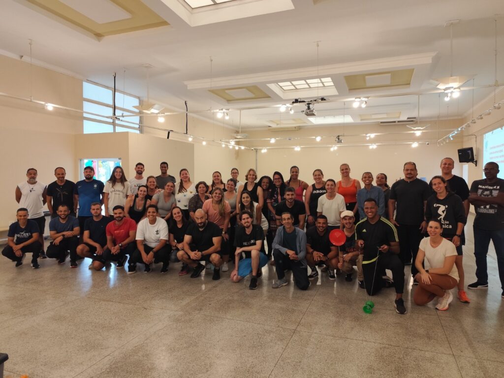 A imagem mostra um grupo grande de pessoas, cerca de 45 adultos, reunidas dentro de um salão espaçoso e bem iluminado. O grupo está posicionado em três fileiras: a primeira fila está agachada ou sentada no chão, enquanto as duas fileiras atrás estão em pé. A maioria está vestida com roupas esportivas ou casuais, como camisetas, regatas, shorts e leggings. Algumas marcas conhecidas aparecem nas roupas, como Adidas, Hollister e Nike. O ambiente é moderno, com teto alto e iluminação de lâmpadas embutidas, além de janelas altas que deixam entrar luz natural. O piso é de pedra ou cerâmica cinza. À direita, um homem está segurando um ioiô verde e um prato plástico vermelho. Todos parecem felizes e sorridentes, sugerindo que estão participando de um evento coletivo, possivelmente uma atividade física, oficina ou treinamento em grupo. Ao fundo, há uma tela de projeção com texto, e uma caixa de som está instalada na parede, indicando que o local está preparado para apresentações ou palestras. A atmosfera geral é de descontração e colaboração.