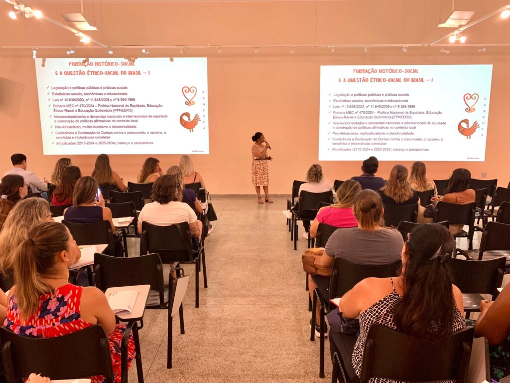 A imagem mostra uma sala de aula ou auditório onde uma mulher está fazendo uma apresentação para um grupo de pessoas sentadas em cadeiras organizadas em fileiras. A apresentadora está em pé, segurando um microfone, e usa um vestido laranja com estampas brancas. À frente dela, há duas telas grandes exibindo o mesmo slide, que está em português e tem o título "FORMAÇÃO HISTÓRICO-SOCIAL E A QUESTÃO ÉTNICO-RACIAL NO BRASIL - I". O slide aborda temas como legislação, políticas públicas, estatísticas e conceitos relacionados à questão étnico-racial no Brasil, mencionando leis específicas, políticas educacionais e movimentos como o Pan-Africanismo e a decolonialidade. A sala é bem iluminada, com luzes no teto e paredes claras. O público, composto principalmente por mulheres, está atento à apresentação, algumas fazendo anotações. No rodapé do slide aparece o nome "Marilda Soares" e a palavra "SANKOFA", que é um símbolo africano representado por um pássaro, visível ao lado do texto. A atmosfera é formal e educativa, indicando um evento de formação ou palestra voltada para temas sociais e étnicos.
