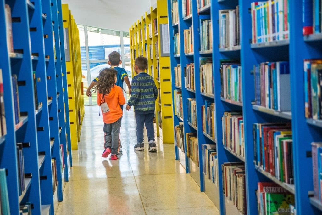 A imagem mostra três crianças caminhando por um corredor de uma biblioteca. Elas estão de costas para a câmera, avançando em direção a uma grande janela ao fundo que deixa a luz natural entrar, iluminando todo o ambiente. À esquerda, há estantes amarelas e, à direita, estantes azuis, ambas repletas de livros. O chão é claro e refletivo, e a composição da foto utiliza uma perspectiva simétrica que destaca as linhas das estantes convergindo para a janela no final do corredor. A criança à esquerda veste um moletom laranja, a do meio uma camiseta azul e amarela, e a da direita uma camisa azul com estampa. O ambiente transmite uma atmosfera de aprendizado e curiosidade, típica de uma biblioteca escolar ou pública.