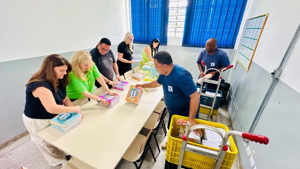 A imagem mostra um grupo de sete pessoas organizadas em torno de uma longa mesa clara, em um ambiente que parece ser uma escola ou um escritório administrativo. Elas estão distribuindo pilhas de livros didáticos, alguns embalados em plástico. À esquerda, duas mulheres e um homem estão organizando os livros; uma mulher veste uma blusa preta e calça branca, a outra uma camiseta verde-limão, e o homem está com camiseta escura e óculos. No centro, um homem com camiseta azul com logo está colocando uma pilha de livros de capa laranja sobre a mesa. Ao lado dele, outro homem, também com camiseta azul, está descarregando livros de um carrinho. Ao fundo, duas mulheres estão analisando papéis e livros. O ambiente tem paredes brancas com uma faixa cinza na parte inferior, janelas com persianas azuis, e um quadro branco com uma grade. No chão, há caixas plásticas amarelas e azuis cheias de livros. A iluminação é clara e uniforme, indicando que a foto foi tirada durante o dia. A cena transmite uma sensação de trabalho colaborativo e organização, provavelmente para preparar materiais escolares.