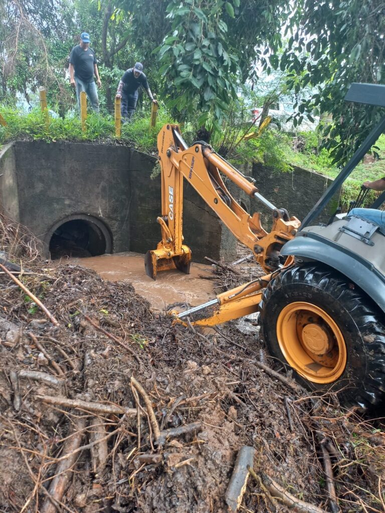 A imagem mostra uma retroescavadeira amarela da marca CASE, com a inscrição "BRASIF" em seu braço, operando para limpar uma área de drenagem. A máquina está posicionada próxima a uma estrutura de concreto que possui uma grande tubulação circular pela qual a água provavelmente escoa. A retroescavadeira está removendo lama e detritos acumulados em um ponto onde a água está bastante turva e barrenta. No primeiro plano, há uma grande quantidade de lama úmida e galhos acumulados, provavelmente retirados do local para desobstruir o fluxo de água. Ao fundo, há vegetação verde e algumas árvores, além de duas pessoas observando o trabalho, uma vestindo camiseta preta e boné azul, e outra com camisa escura e boné. À direita, é possível ver parcialmente a mão e a perna do operador da máquina. O cenário sugere que o trabalho está sendo realizado para desobstruir a passagem da água, possivelmente após uma chuva forte ou enchente, garantindo a correta drenagem e evitando alagamentos. O ambiente é externo, provavelmente em uma área rural ou de mata.