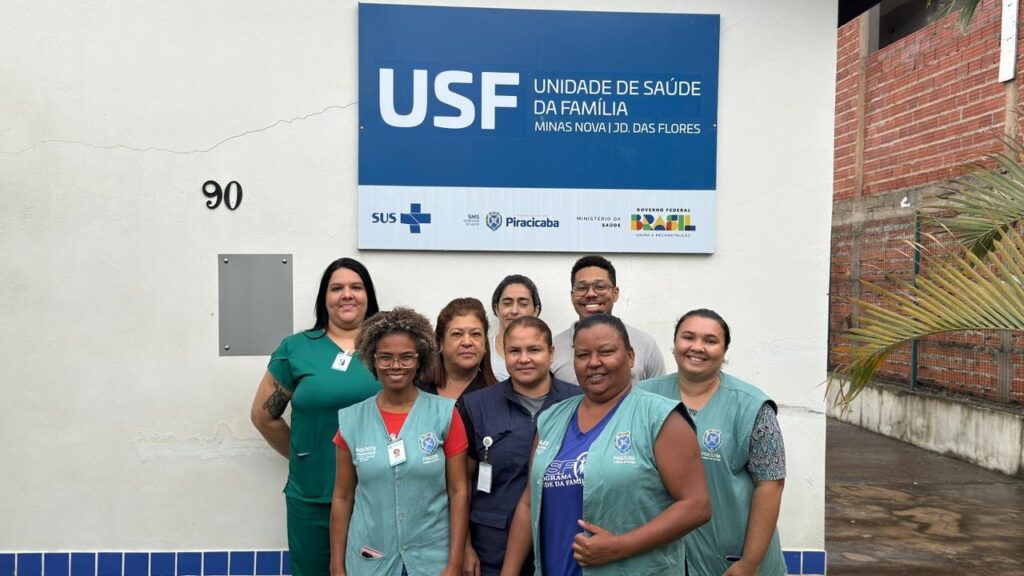 A imagem mostra um grupo de oito profissionais de saúde posando para uma foto em frente a uma unidade de saúde. Eles estão alinhados lado a lado, todos sorrindo para a câmera. A maioria veste uniformes médicos, como jalecos verdes e coletes azuis, e alguns usam crachás de identificação. Ao fundo, há uma parede branca com uma placa azul e branca que indica o nome da unidade: "USF Unidade de Saúde da Família Minas Nova | JD. das Flores". Também estão visíveis os logos do SUS, Secretaria de Saúde de Piracicaba, Ministério da Saúde e Governo Federal do Brasil. À esquerda da imagem, está o número "90", provavelmente o número do prédio ou endereço. A cena parece ser ao ar livre, com iluminação natural e um ambiente limpo e organizado, transmitindo um clima de profissionalismo e acolhimento.
