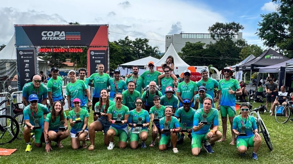 A imagem mostra uma grande equipe de atletas reunidos ao ar livre, posando para uma foto em grupo durante um evento esportivo chamado "COPA INTERIOR". São cerca de 25 pessoas, homens e mulheres, vestindo uniformes esportivos nas cores verde e azul, com o logo "care club". Muitos seguram pequenos troféus, indicando que participaram e possivelmente venceram em alguma competição. O cenário é um campo gramado, com uma estrutura preta e vermelha ao fundo que exibe um cronômetro digital com os números "04:03:41", "032024" e "02:33:46". Também há tendas brancas e pretas com marcas como "FIKFIT" e outras, além de bicicletas espalhadas ao redor. O clima parece ser de dia, com luz natural e céu levemente nublado. A imagem transmite um ambiente de celebração esportiva, união da equipe e realização após uma competição.