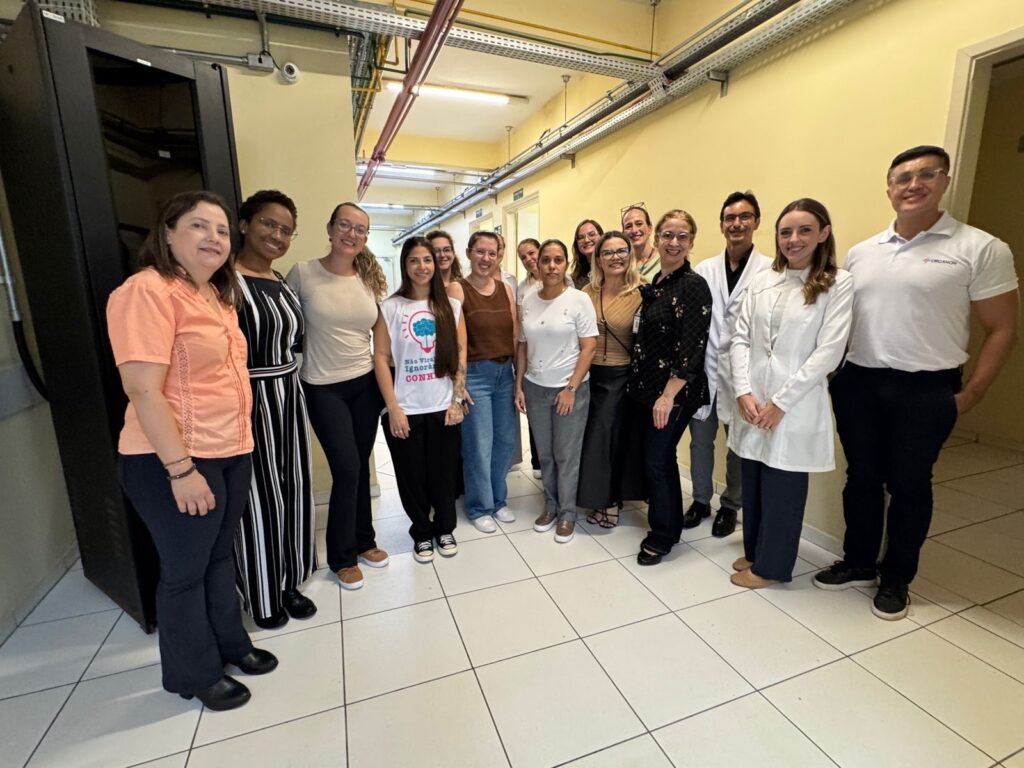 A imagem mostra um grupo de 15 pessoas, em sua maioria mulheres, posando para uma foto em um corredor interno estreito e bem iluminado. Elas estão organizadas em duas fileiras, todas sorrindo para a câmera. O ambiente parece ser profissional, possivelmente relacionado à área da saúde ou ciência, pois algumas pessoas vestem jalecos brancos e roupas de scrubs. O corredor tem paredes amarelas claras e piso de azulejos brancos com rejunte escuro. Do lado esquerdo, há um grande armário ou rack, possivelmente de equipamentos técnicos. No teto, tubulações e cabos estão aparentes. As roupas das pessoas variam entre casual e profissional, incluindo camisetas, jeans, jalecos e uniformes. O clima é amistoso e colaborativo. No centro, uma mulher usa uma camiseta branca com a imagem de um cérebro e texto em português, e no canto direito, um homem veste uma camisa polo branca com um logo vermelho. No fundo, há palavras parciais visíveis nas paredes, como "Não Vira", "Ignorâ", "CONHE" e "ORGANON", indicando que o local pode ser uma instituição ou campanha relacionada à conscientização ou conhecimento.