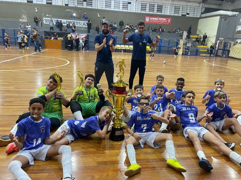 A imagem mostra um grupo de jovens jogadores de futsal, acompanhados por dois treinadores adultos, comemorando uma vitória dentro de um ginásio esportivo. No centro da cena, destaca-se um troféu grande e dourado, que simboliza o prêmio conquistado pela equipe. Os jogadores estão vestidos com uniformes azuis e shorts brancos, alguns usando medalhas e óculos escuros, demonstrando alegria e descontração. Dois jogadores, provavelmente goleiros, vestem uniformes verdes e seguram troféus menores. Os treinadores, posicionados atrás dos atletas, estão com os braços levantados em sinal de celebração. Ao fundo, é possível ver arquibancadas com algumas pessoas assistindo e banners que indicam que o evento está relacionado ao "Atibaia Futsal" e à "Secretaria de Esportes e Lazer". O ambiente é iluminado, com o piso de madeira típico de quadras esportivas. A atmosfera geral da imagem é de vitória, união e entusiasmo juvenil.