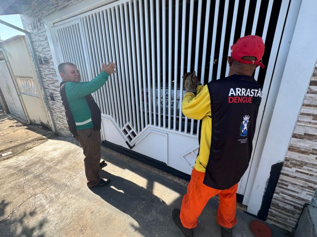 A imagem mostra dois homens em frente a um portão branco de uma residência durante o dia. Um dos homens está de perfil, vestindo uma camisa verde com colete escuro e calça marrom, e parece estar gesticulando ou batendo palmas em direção ao portão. O outro homem está de costas, usando um boné vermelho, camisa amarela com colete preto e calça laranja. No colete dele está escrito "ARRASTÃO DENGUE 202" e "A SERVIÇO DA PREFEITURA MUNICIPAL DE PIRACICABA", indicando que ele é um agente de saúde pública. Ao fundo, atrás do portão, é possível ver parte de um carro prata estacionado. A casa tem uma parede com textura que imita pedras e o número 1075 está visível na parede. A cena parece ser parte de uma ação de combate ao mosquito da dengue, realizada pela prefeitura da cidade de Piracicaba. A iluminação é forte, com sombras bem definidas no chão.