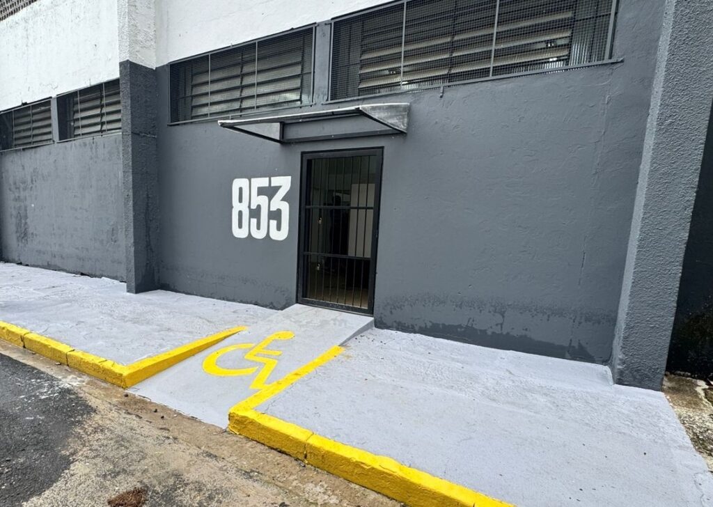A imagem mostra a fachada de um edifício urbano com uma porta de metal preta, protegida por uma grade também preta. Ao lado da porta, está pintado em branco o número "853". Acima da porta, há uma pequena cobertura metálica e janelas com telas escuras. Na calçada em frente, há uma rampa de acesso para pessoas com deficiência, destacada pela pintura amarela com o símbolo internacional de acessibilidade (cadeirante). A calçada é de concreto cinza claro, e o meio-fio é pintado de amarelo, indicando a área para acesso facilitado. A iluminação é natural, sugerindo que a foto foi tirada durante o dia. A cena transmite uma sensação de funcionalidade e acessibilidade no ambiente urbano.