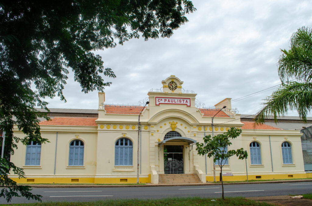 A imagem mostra a fachada histórica da Companhia Paulista de Estradas de Ferro, um edifício clássico e imponente. A construção é de cor creme com detalhes arquitetônicos em estilo neoclássico, incluindo janelas simétricas com persianas azuis e telhados de telha laranja. No centro da fachada há um grande arco com um portão de ferro trabalhado, acima do qual está uma placa vermelha com a inscrição "CIA PAULISTA" em letras brancas. No topo do edifício, há uma torre com um relógio, que reforça o caráter antigo e tradicional do prédio. Em primeiro plano, há algumas árvores verdes que enquadram a cena, e o céu está nublado, criando uma iluminação suave e difusa que destaca os detalhes da construção. A imagem transmite uma sensação de história e importância, típica de uma antiga estação ferroviária ou sede administrativa de uma companhia ferroviária.