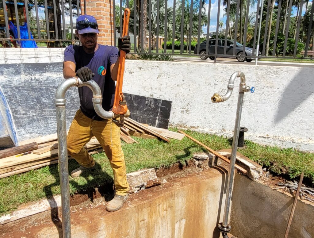 A imagem mostra um encanador trabalhando ao ar livre em uma instalação de tubos. Ele está dentro de uma escavação, usando uma camisa azul, calças de trabalho bege, boné azul com óculos escuros sobre ele e luvas pretas. O homem está segurando uma chave de grifo laranja grande e está ajustando um tubo metálico vertical que possui uma curva em forma de "U" na parte superior. Ao redor, há tábuas de madeira sobre a grama e, ao fundo, um carro preto estacionado próximo a uma cerca metálica preta e uma parede branca. O ambiente parece ser um local de manutenção ou construção, com iluminação natural forte, indicando que é durante o dia.