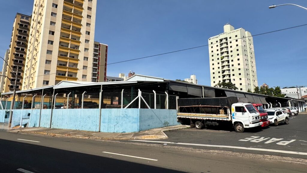 A imagem mostra uma cena urbana durante um dia claro e ensolarado. Em primeiro plano, há uma rua asfaltada com a palavra "PARE" pintada no chão, indicando uma parada obrigatória para os veículos. À direita, estão estacionados vários veículos, incluindo caminhões e carros. No centro da imagem, destaca-se um prédio baixo, possivelmente um galpão ou depósito, com uma base azul clara e uma cerca de arame no topo. Ao fundo, erguem-se prédios residenciais altos, de diferentes estilos e cores, como bege com detalhes amarelos e um tom claro uniforme. O céu está azul e sem nuvens, e a iluminação forte sugere que a foto foi tirada ao meio-dia. A cena transmite uma mistura de áreas industriais e residenciais em um ambiente urbano.