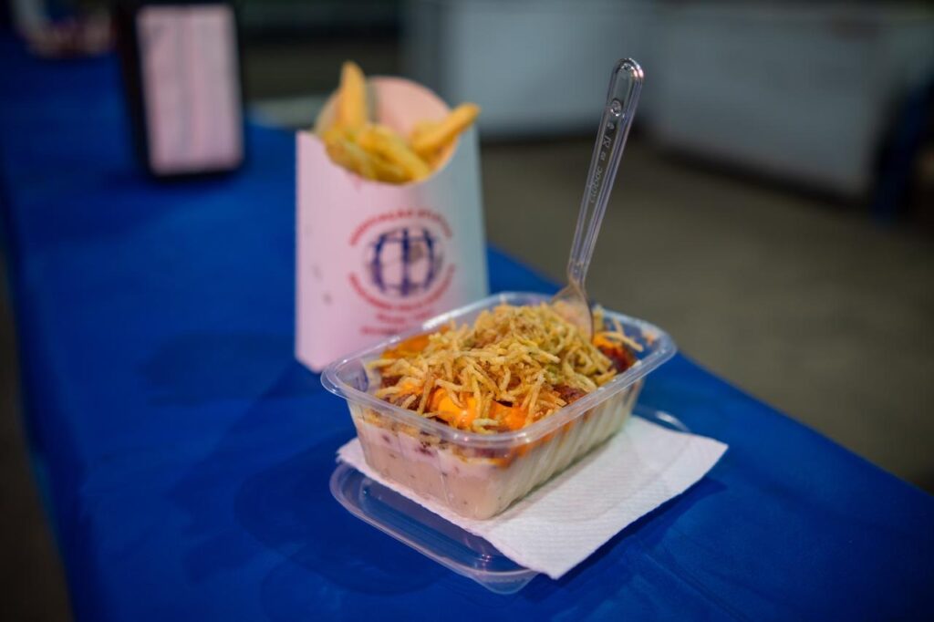 A imagem mostra uma porção de comida servida em um recipiente plástico transparente, contendo uma mistura de ingredientes que parecem ser carne, queijo e batata palha por cima. Ao lado, há um cone de papel branco com batatas fritas dentro, apoiado sobre uma toalha de mesa azul. Um garfo plástico transparente está fincado na comida. O fundo está desfocado, destacando o prato principal. A cena parece estar em um ambiente informal, como uma feira ou evento de comida. A combinação sugere um prato típico de comida rápida ou de rua, possivelmente uma versão de cachorro-quente ou um prato brasileiro conhecido por ingredientes similares.