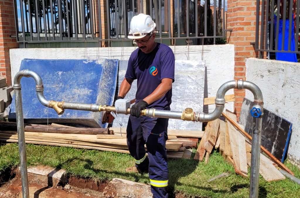 A imagem mostra um trabalhador masculino realizando a instalação ou manutenção de um conjunto de tubulações metálicas ao ar livre. Ele está usando um capacete branco com o logotipo "SANE SOLUTIONS", óculos de proteção, luvas pretas, camiseta azul escura com um símbolo estilizado e calças de trabalho com faixas reflexivas amarelas e a inscrição "SANESOLUTI". O trabalhador segura firmemente uma tubulação horizontal de metal galvanizado, que está conectada a válvulas de latão e a uma pequena caixa eletrônica branca com o número "20". O ambiente é um canteiro de obras ou local de infraestrutura, com uma parede de tijolos e uma cerca de metal ao fundo, além de vegetação e materiais de construção ao redor. A cena ocorre durante o dia, com luz natural forte e sombras definidas. O conjunto sugere trabalho relacionado a sistemas de água ou saneamento.