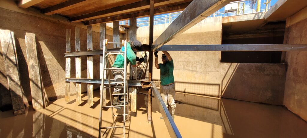 A imagem mostra um ambiente industrial, possivelmente uma grande cisterna ou reservatório de concreto, com paredes altas e robustas. Dentro desse espaço, há dois trabalhadores realizando uma tarefa de manutenção ou construção. Um dos trabalhadores está sobre uma escada de alumínio do tipo escada A, vestindo camiseta verde, calça cinza e botas pretas, e parece estar fixando ou ajustando uma viga de metal galvanizado. O outro trabalhador está dentro da água turva que cobre o fundo da estrutura, usando camiseta escura e calça clara suja de lama, auxiliando na instalação das vigas metálicas. Ao redor deles, vê-se uma estrutura de vigas metálicas fixadas nas paredes de concreto, com suportes de madeira verticais. A iluminação é natural, vinda de cima, indicando que a estrutura está aberta no topo, e o céu azul é visível acima das paredes do reservatório. A cena transmite uma sensação de trabalho pesado e cuidado na manutenção de uma grande estrutura industrial.