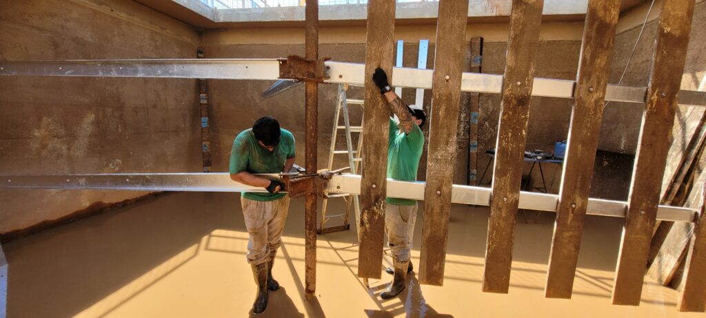 A imagem mostra dois trabalhadores da construção civil dentro de uma estrutura de concreto, possivelmente uma cisterna ou reservatório, parcialmente cheia de água turva e marrom. Ambos vestem camisetas verdes, calças claras e botas pretas de borracha. Um dos trabalhadores está agachado, aparentemente ajustando ou fixando uma peça de metal que faz parte de uma estrutura metálica que está sendo montada dentro do tanque. O outro está em pé, segurando uma tábua de madeira verticalmente, que faz parte de uma série de tábuas alinhadas. A estrutura metálica é composta por vigas horizontais prateadas, sustentadas por um poste vertical enferrujado no centro. Ao fundo, há uma escada de metal dentro da água e uma pequena mesa com ferramentas. As paredes de concreto ao redor mostram sinais de umidade e sujeira, e a luz natural que entra por uma abertura no topo ilumina a cena, destacando os trabalhadores e suas atividades. A imagem transmite uma sensação de trabalho físico intenso em um ambiente úmido e desafiador.