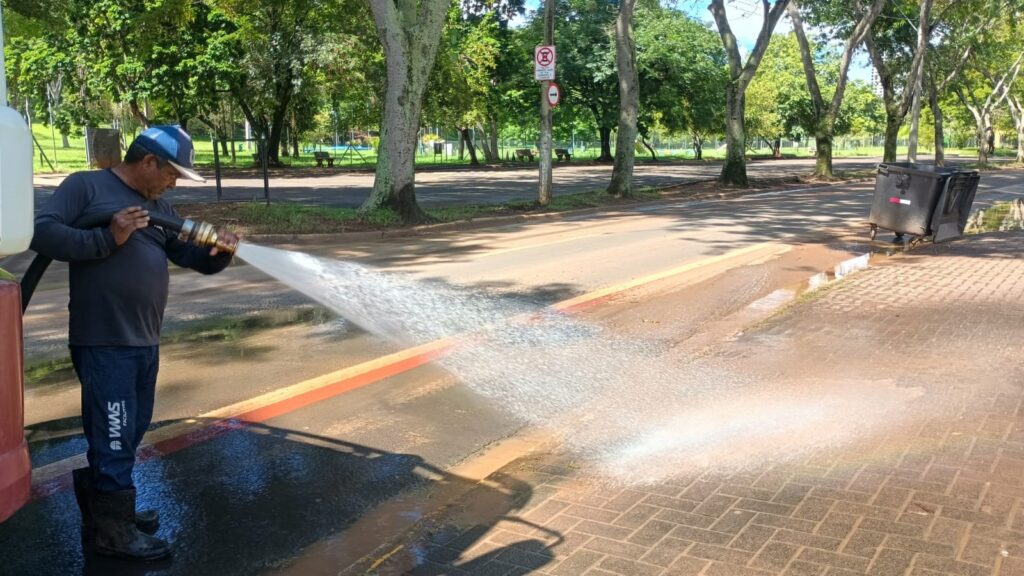 A imagem mostra um trabalhador realizando a limpeza de uma área pavimentada usando uma mangueira de alta pressão. Ele está vestido com uniforme azul, incluindo uma camisa de manga longa, calça escura com o logo "VWS FACILITIES" e um boné azul. O homem está focado na tarefa, direcionando o jato de água para a calçada de tijolos ao lado da rua asfaltada, que tem uma faixa pintada de vermelho. Ao fundo, há várias árvores, indicando que a área é provavelmente um parque ou uma via arborizada. Também é possível ver uma lixeira preta e algumas placas de sinalização, incluindo uma de proibição de estacionamento. A cena ocorre durante o dia, com luz natural forte e sombras longas, sugerindo que seja pela manhã ou fim de tarde.