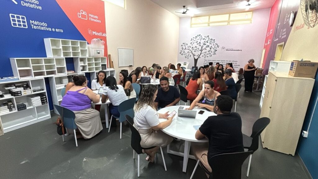 A imagem mostra um ambiente moderno e bem iluminado, provavelmente uma sala de aula ou espaço de treinamento, onde cerca de 20 a 25 pessoas estão distribuídas em grupos ao redor de mesas redondas brancas. A maioria das pessoas parece estar envolvida em discussões ou atividades colaborativas, algumas utilizando laptops e celulares. As paredes possuem uma decoração colorida, com gráficos e textos em português, destacando métodos de aprendizado como "Método Detetive", "Método Tentativa e erro" e "Método Filmadora", além das marcas "Mind Lab" e "MenteInovadora". O ambiente é organizado e acolhedor, com estantes contendo materiais diversos, cadeiras modernas e iluminação natural que entra pelas janelas altas. A atmosfera transmite um clima de aprendizado dinâmico e interativo.