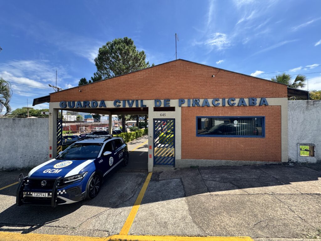 A imagem mostra uma viatura da Guarda Civil Municipal (GCM) de Piracicaba, estacionada em frente a um prédio que parece ser a sede ou um posto da corporação. O veículo é um Volkswagen Nivus na cor azul com detalhes em branco, equipado com sirene e luzes de emergência no teto, além de uma grade protetora na frente. A viatura está posicionada de lado, permitindo ver claramente os detalhes como o emblema da GCM, o número 153 (provavelmente o telefone de emergência) e a placa TJC7F41. O prédio ao fundo é de um pavimento, feito em alvenaria com tijolos aparentes e uma grande placa azul com letras brancas que diz "GUARDA CIVIL DE PIRACICABA". Acima da porta de entrada há o número 645. O local está bem iluminado por luz natural, com céu azul e poucas nuvens, indicando que a foto foi tirada durante o dia. No topo do prédio, há uma antena, sugerindo comunicação via rádio. No geral, a imagem transmite uma sensação de organização e prontidão da Guarda Civil Municipal de Piracicaba para atender à comunidade local.