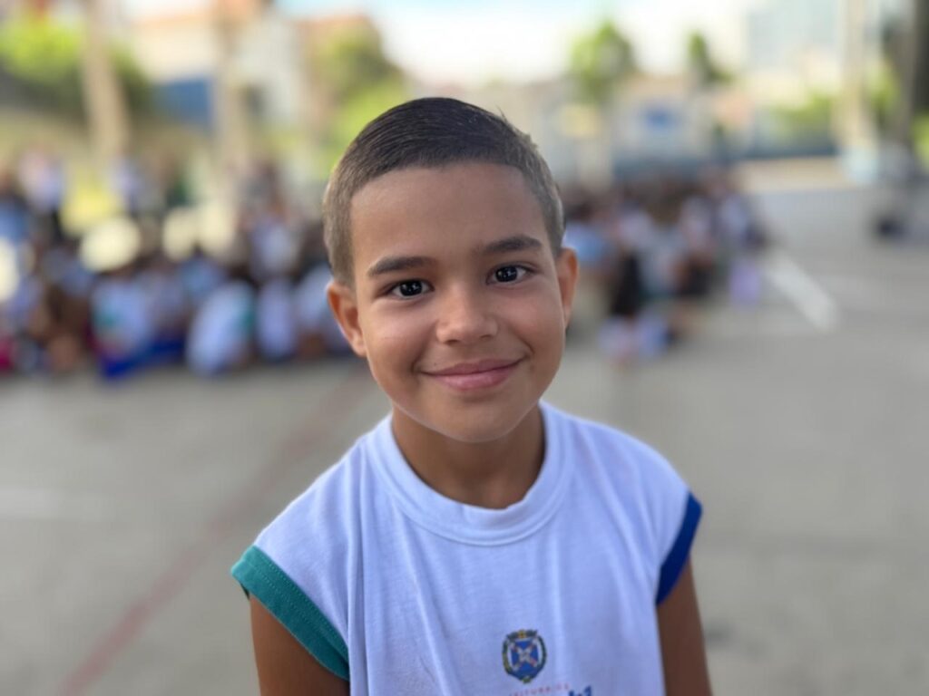 A imagem mostra um menino jovem, provavelmente entre 7 e 10 anos, em destaque no primeiro plano, olhando diretamente para a câmera com um leve sorriso. Ele veste uma camiseta regata branca com detalhes em azul e verde, que possui um brasão ou logo no peito com a palavra "PREFEITURA", indicando que pode ser um uniforme escolar de alguma instituição pública, provavelmente no Brasil, dado o uso do português. Ao fundo, desfocado, há um grupo grande de outras crianças sentadas no chão, sugerindo um ambiente escolar ou um evento comunitário ao ar livre. O cenário é iluminado por luz natural, com o chão de concreto claro e uma atmosfera tranquila e acolhedora. A foto foi tirada em um ângulo ao nível dos olhos do menino, com foco nítido nele e o fundo desfocado, destacando-o como o principal sujeito da imagem.