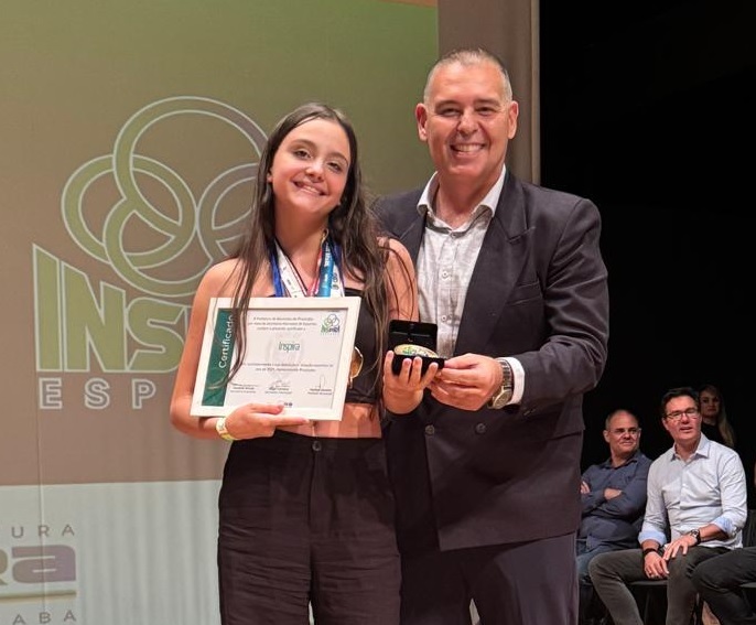 A imagem mostra uma cerimônia de premiação realizada em um palco. No centro, uma jovem sorridente veste uma blusa preta curta e calças pretas, usando várias medalhas no pescoço e segurando um certificado emoldurado. Ao lado dela, um homem mais velho, também sorridente, está vestido com um paletó escuro e camisa clara, segurando uma caixa aberta com uma medalha dentro. Ao fundo, há um banner grande com a palavra "INSPIRA" e um logotipo com círculos entrelaçados. Três homens estão sentados ao fundo, observando a cerimônia. A iluminação é clara, destacando os premiados, e o ambiente transmite uma atmosfera de celebração e reconhecimento.