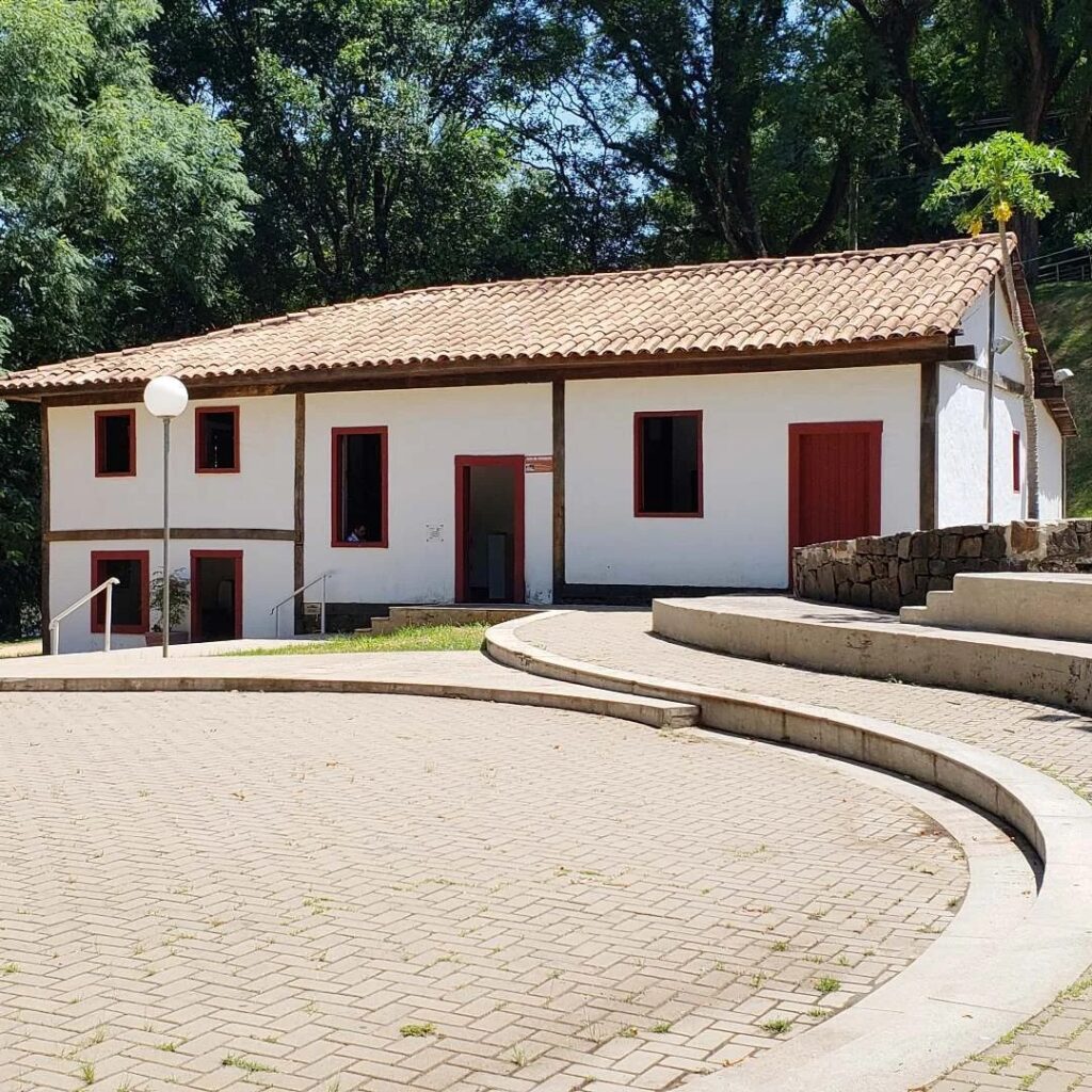 A imagem mostra um edifício colonial de um andar, com paredes brancas e estrutura de madeira escura aparente, típico da arquitetura colonial. O telhado é de telhas cerâmicas vermelhas em estilo colonial. As portas e janelas são pintadas em vermelho escuro. O prédio está localizado em um terreno levemente inclinado, com uma parte inferior visível à esquerda, onde há duas janelas pequenas e uma porta. Em frente ao edifício há uma ampla praça pavimentada com pedras claras, com degraus curvos que parecem servir como bancos ou áreas de descanso. Ao redor, há vegetação verde densa, incluindo uma árvore alta que parece ser de mamão, e uma parede de pedra ao lado direito da imagem. A iluminação é natural, com luz solar forte que cria sombras definidas, sugerindo que a foto foi tirada durante o dia, em um ambiente tranquilo e bem cuidado. A cena transmite uma atmosfera histórica e cultural, possivelmente de uma construção preservada em uma região tropical ou subtropical.