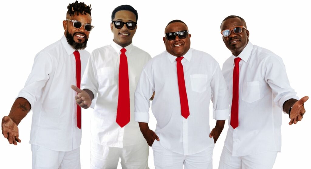 A imagem mostra quatro homens negros alinhados lado a lado, todos vestindo camisas sociais brancas de manga longa, calças brancas e gravatas vermelhas vibrantes. Cada um usa um tipo diferente de óculos escuros. Eles estão contra um fundo branco liso, com iluminação forte e uniforme, que destaca suas roupas e expressões. Os homens parecem alegres e confiantes, com gestos amigáveis, transmitindo uma sensação de unidade e estilo. A imagem tem um ar profissional e pode sugerir que eles fazem parte de um grupo musical ou equipe coordenada.