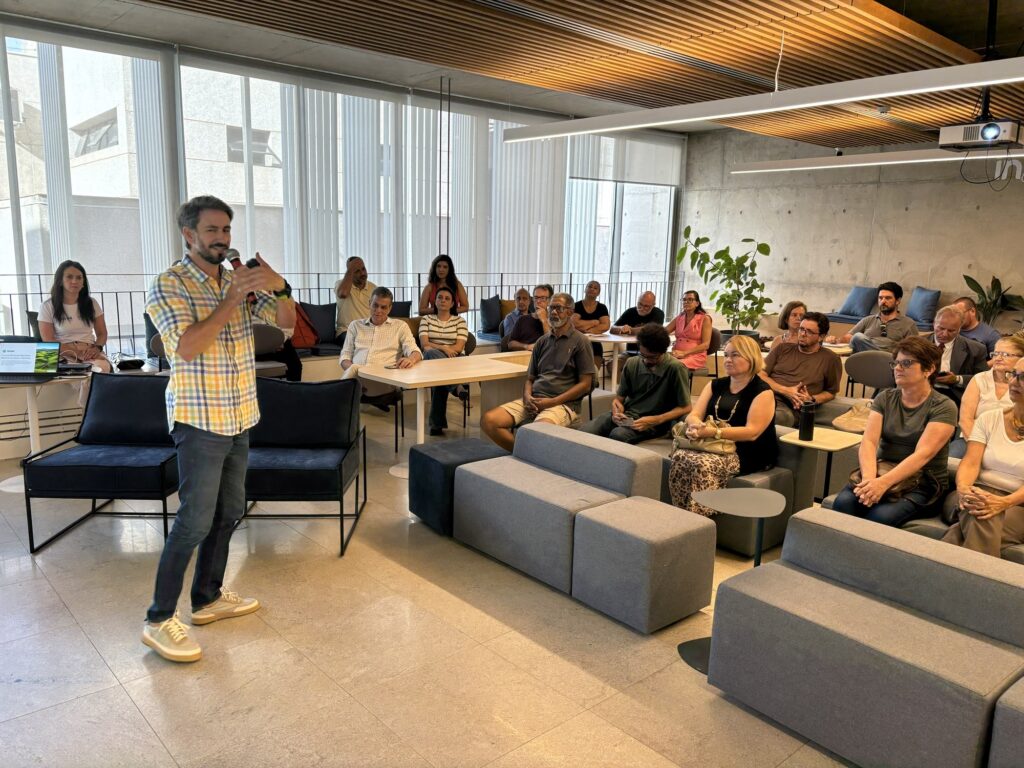 A imagem mostra uma apresentação ou palestra acontecendo em um ambiente interno moderno e bem iluminado, com grandes janelas que permitem a entrada de luz natural. Um homem, vestido com uma camisa xadrez amarela e azul, está em pé segurando um microfone, falando para um grupo de pessoas sentadas em pufes e cadeiras. O público, composto por cerca de 20 pessoas de diferentes idades e etnias, está atento ao palestrante. O espaço é decorado com móveis modernos, incluindo um sofá preto, pufes cinza e mesas de madeira clara. No teto, há painéis de madeira e um projetor está instalado, sugerindo que pode haver uma apresentação visual. Um laptop próximo ao palestrante exibe o texto "SENAR Turismo Rural em São Paulo História de valores e natureza", indicando que o tema da palestra está relacionado ao turismo rural no estado de São Paulo. O ambiente parece ser um local de trabalho colaborativo ou uma sala de eventos, com uma parede de concreto ao fundo e a logo do LinkedIn visível em uma das paredes. A atmosfera é profissional e acolhedora, com pessoas engajadas na discussão ou apresentação.