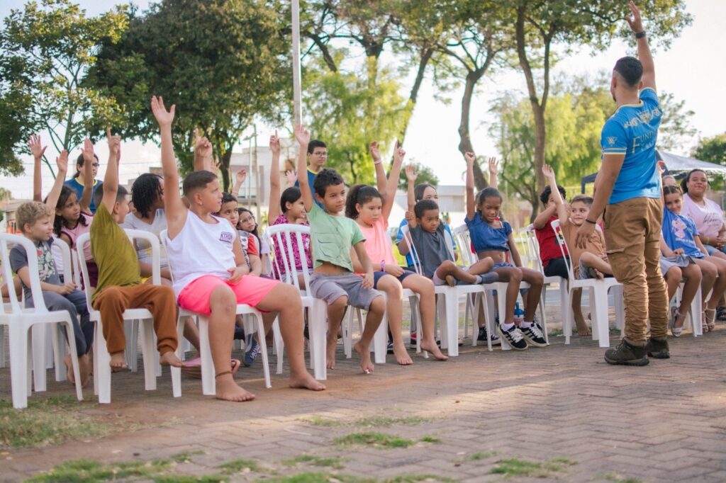 A imagem mostra um grupo de crianças sentadas em cadeiras plásticas brancas em um ambiente externo, provavelmente um parque ou área comunitária. As crianças estão organizadas em filas e muitas delas estão com as mãos levantadas, indicando que estão participando ativamente de uma atividade ou respondendo a uma pergunta. À direita, um homem adulto, possivelmente o instrutor ou professor, também está com a mão levantada, interagindo com o grupo. Ele veste uma camiseta azul com a palavra "FUND" escrita em dourado. O cenário é iluminado pela luz natural do dia, com árvores verdes ao fundo, criando um ambiente agradável e ao ar livre. A cena transmite uma atmosfera de aprendizado, diversão e engajamento comunitário.
