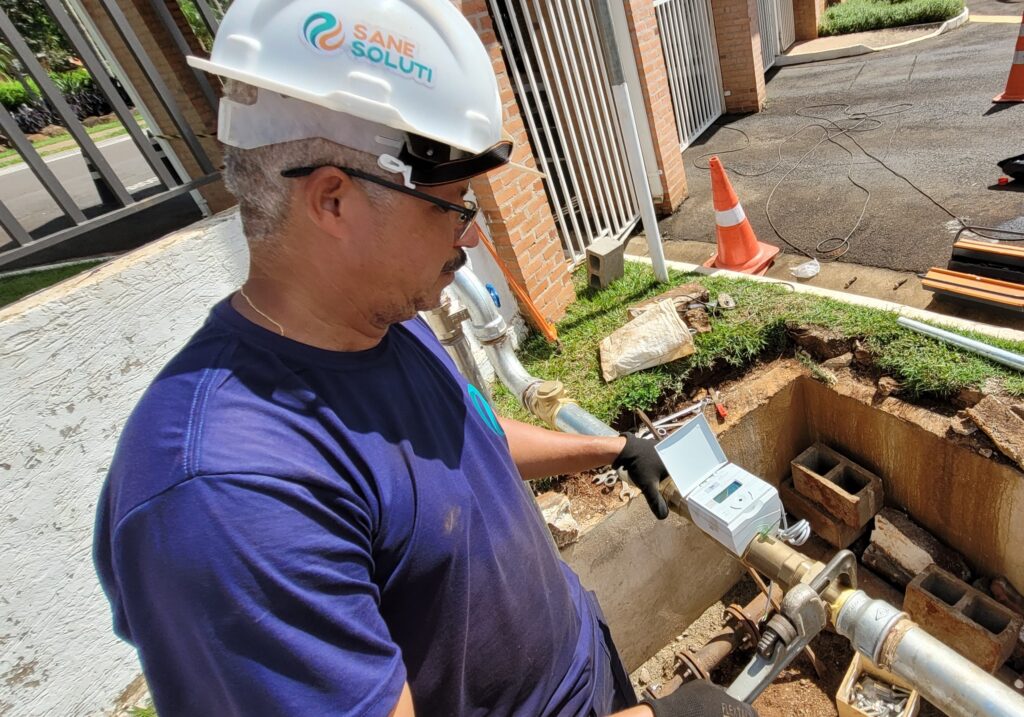 A imagem mostra um trabalhador masculino, que está usando capacete branco e óculos de proteção, realizando a instalação ou inspeção de um hidrômetro em uma abertura no chão. Ele veste uma camiseta azul e luvas pretas, e parece estar concentrado na tarefa. O hidrômetro é branco com um visor digital, conectado a tubos metálicos dentro da abertura. Ao redor do local, há cones de sinalização laranja e branco, blocos de concreto e uma parede de tijolos ao fundo, indicando que o serviço está sendo feito em uma área externa, possivelmente residencial ou comercial. O capacete do trabalhador possui o logo "SANE SOLUTI", que sugere que ele trabalha para uma empresa relacionada a saneamento ou serviços públicos de água. A imagem foi capturada em um dia claro, com luz natural forte iluminando a cena.