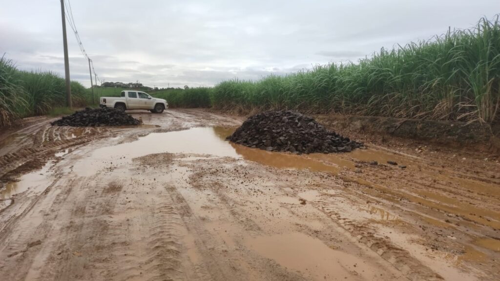 A imagem mostra uma estrada rural de terra, bastante lamacenta e cheia de poças d'água, provavelmente após uma chuva. No centro da estrada, há duas grandes pilhas de pedras escuras, possivelmente basalto, que parecem estar ali para ajudar na manutenção ou para melhorar a passagem naquele trecho. Ao lado da estrada, há plantações de cana-de-açúcar com folhas verdes e altas, indicando que se trata de uma área agrícola. Do lado esquerdo, há postes de energia elétrica alinhados ao longo da estrada. Ao fundo, um caminhonete branca está estacionada na lateral da estrada. O céu está nublado, com nuvens cinzentas, o que reforça a impressão de tempo chuvoso e ambiente úmido. A estrada se estende em direção ao horizonte, levemente curvada para a esquerda. A cena transmite uma sensação de um local rural simples, em meio à natureza e à atividade agrícola.