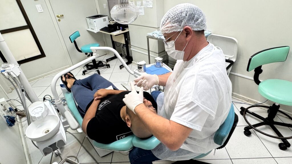 A imagem mostra um consultório odontológico durante um atendimento. Um dentista, vestido com touca, máscara, óculos, jaleco branco e luvas, está inclinado sobre um paciente que está reclinado em uma cadeira odontológica azul-turquesa. O paciente é um homem com cabeça raspada, usando camiseta preta e calça azul, e parece estar recebendo um procedimento dental. Ao redor, é possível ver equipamentos odontológicos, como uma pia para enxágue, uma luminária odontológica e uma cadeira adicional ao fundo. O ambiente é iluminado e limpo, com paredes claras e piso de azulejo. Há também uma impressora sobre uma mesa próxima à porta fechada. A imagem transmite uma cena típica de atendimento odontológico em um ambiente profissional.