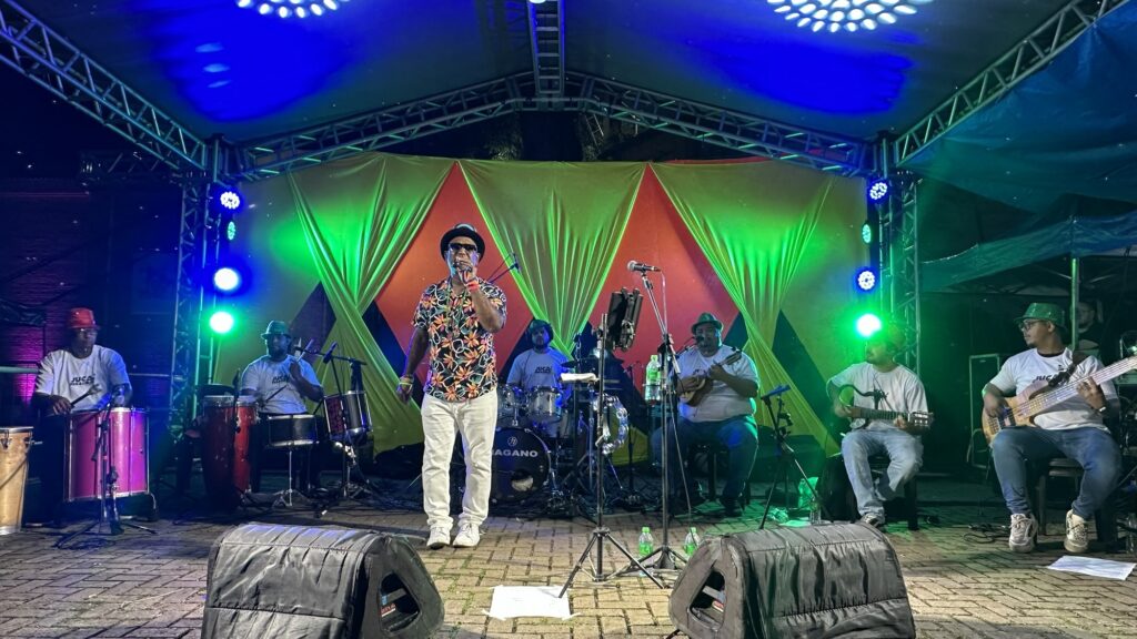 A imagem mostra uma apresentação musical ao ar livre durante a noite. No centro do palco, há um cantor principal usando um chapéu preto estilo fedora, óculos escuros, uma camisa floral colorida e calças brancas, segurando um microfone. Ao redor dele, estão os músicos da banda: três percussionistas à esquerda, um deles tocando conga, outro em um pequeno conjunto de tambores, e um terceiro em um grande tambor rosa; à direita do cantor, há um baterista atrás de uma bateria com a marca "NAGANO" e dois guitarristas sentados, um tocando guitarra acústica e outro o contrabaixo. Todos os músicos, exceto o cantor principal, vestem camisetas brancas com um logotipo e chapéus verdes. O palco é decorado com tecidos amarelos, verdes e vermelhos ao fundo, e estruturas metálicas sustentam as luzes coloridas que iluminam o ambiente em tons de azul e verde. No chão, à frente do palco, estão dois alto-falantes grandes voltados para o público. A atmosfera é vibrante e festiva, típica de um evento musical ao vivo.