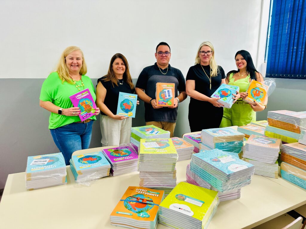 A imagem mostra cinco adultos, três mulheres e dois homens, posicionados atrás de uma mesa longa repleta de pilhas de livros coloridos. Todos estão sorrindo e segurando livros individuais voltados para a câmera. Da esquerda para a direita, há uma mulher vestindo uma camiseta verde neon e jeans, segurando um livro rosa; uma mulher com camiseta preta e calça branca, segurando um livro azul claro; um homem com camisa polo preta, segurando um livro laranja; uma mulher com vestido preto e óculos, segurando uma pilha de livros azuis; e uma mulher vestindo roupa verde-limão, segurando um livro laranja. Os livros têm o título "LIVRO DO ESTUDANTE" e apresentam na capa imagens de animais como onças, capivaras e preguiças. Sobre a mesa, há vários pacotes plásticos contendo esses livros. O ambiente é interno, com paredes brancas e uma faixa cinza na parte inferior, além de uma janela com persianas azuis à direita. A iluminação é uniforme e clara, típica de um ambiente de escritório ou sala de aula. Na capa dos livros, também aparecem os logos do governo do estado de São Paulo e da Secretaria da Educação.