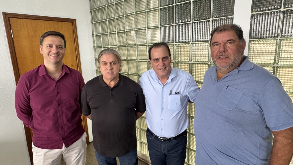 A imagem mostra quatro homens adultos, aparentemente em um ambiente interno, possivelmente um escritório ou local de trabalho. Eles estão posicionados lado a lado, olhando para a câmera e sorrindo de forma amigável. O homem à esquerda veste uma camisa social vinho e calça clara, tem cabelo curto e escuro. O segundo homem usa uma camisa polo preta e jeans, com cabelo grisalho. O terceiro homem veste uma camisa social azul clara, calça jeans escura e usa um cinto; ele também tem uma caneta no bolso da camisa. O quarto homem, à direita, é mais robusto, tem cabelo e barba grisalhos e veste uma camisa polo azul claro. O fundo é simples, com uma parede branca e uma divisória feita de blocos de vidro translúcidos. A iluminação é clara e uniforme, destacando bem as pessoas na foto. A imagem transmite um clima informal, porém profissional.