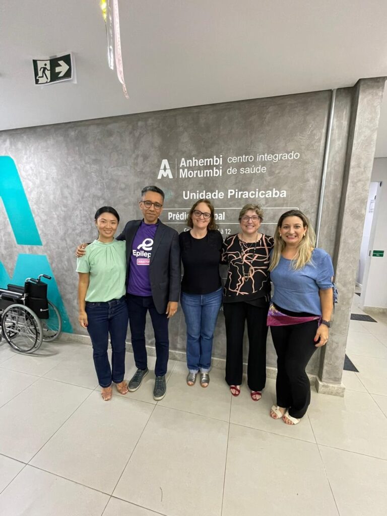 A imagem mostra cinco pessoas alinhadas lado a lado, posando para uma foto em um ambiente interno que parece ser uma unidade de saúde. Atrás deles, há uma parede cinza com o nome “Anhembi Morumbi centro integrado de saúde” e “Unidade Piracicaba” escrito em letras brancas. À esquerda, há uma cadeira de rodas manual encostada na parede, que tem uma pintura ou adesivo em tom verde-azulado. No canto superior esquerdo, há uma placa de saída de emergência com um símbolo de uma pessoa correndo e uma seta. As pessoas na foto estão sorrindo, vestindo roupas casuais e semi-formais. Uma delas usa uma camiseta roxa com um símbolo e a palavra “Epilep”, possivelmente relacionada à epilepsia, indicando que o grupo pode estar envolvido com algum evento ou trabalho relacionado à saúde, mais especificamente à neurologia ou epilepsia. O ambiente é bem iluminado, com piso claro e paredes lisas, transmitindo uma atmosfera profissional e acolhedora, típica de clínicas ou centros de saúde.