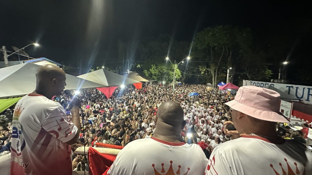 A imagem mostra uma festa de rua noturna muito animada e cheia de pessoas, provavelmente durante um evento cultural ou de Carnaval no Brasil. Na parte frontal da foto, três homens estão de costas ou de lado, sobre uma plataforma ou palco, todos vestidos com camisas brancas com detalhes em vermelho, indicando que fazem parte de um grupo ou escola de samba. Um deles usa um chapéu rosa, enquanto os outros dois são carecas. À frente deles, uma multidão enorme veste roupas similares, em branco e vermelho, e ocupa toda a rua, demonstrando grande entusiasmo e energia. Luzes fortes iluminam a cena, com reflexos e flare, realçando o clima festivo. Ao fundo, é possível ver árvores, algumas barracas brancas e uma placa com a inscrição "TORRES TUR". A imagem transmite a sensação de uma celebração coletiva, com música, dança e muita participação popular, típica das grandes festas brasileiras.