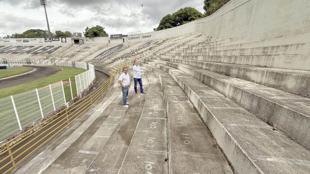 A imagem mostra dois homens em pé nas arquibancadas de concreto de um estádio esportivo ao ar livre, provavelmente no Brasil, dado o idioma das placas e propagandas visíveis. Eles estão vestidos de forma casual, com camisas brancas e calças jeans. Um dos homens está com as mãos na cintura, enquanto o outro aponta em direção ao campo. O estádio está praticamente vazio, com fileiras de assentos de concreto numerados (101 a 110 visíveis). Há uma cerca branca metálica e um corrimão amarelo à frente dos homens. O campo ao lado é verde e cercado por uma pista de atletismo vermelha. No fundo, há várias árvores e o céu está nublado, o que cria uma iluminação difusa e uniforme. Ao redor do estádio, há vários painéis de publicidade com marcas como Uniodonto, Unimed, Raízen, Savegnago, entre outras. O ambiente sugere que os homens podem estar inspecionando ou discutindo algo relacionado ao estádio ou ao evento esportivo que ocorre ali.