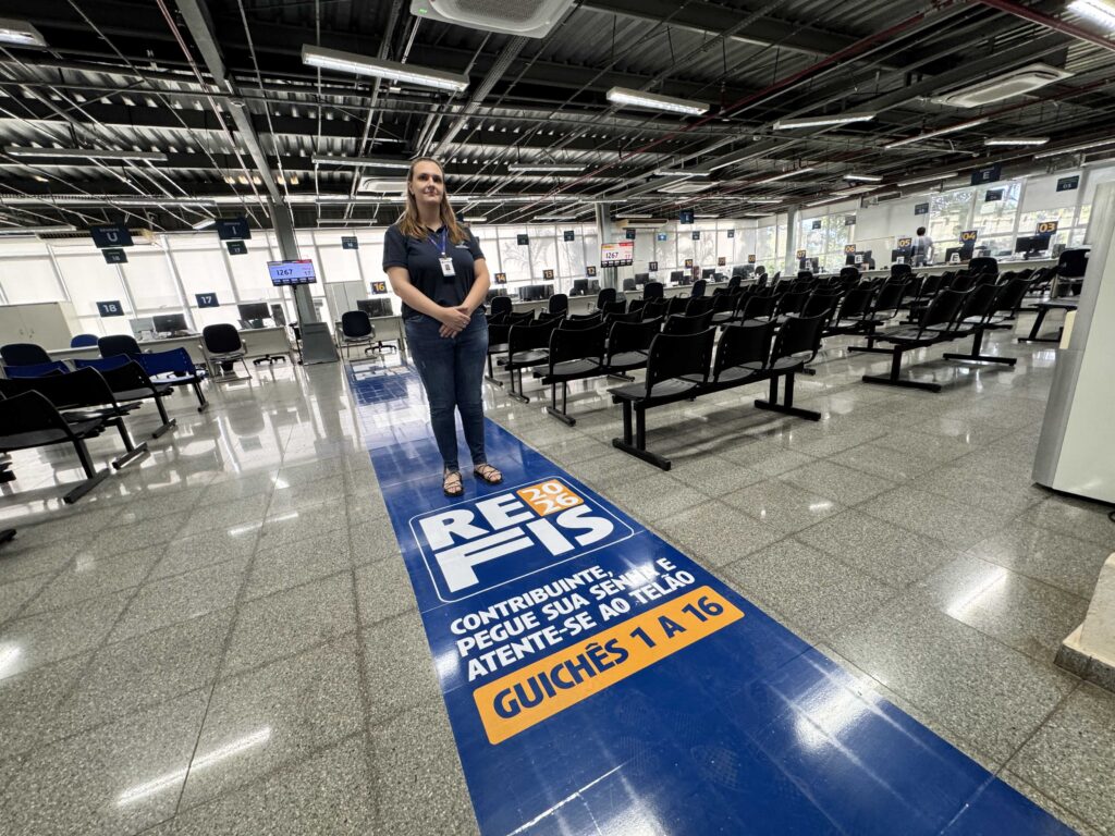 A imagem mostra o interior de um grande salão de atendimento, possivelmente uma repartição pública ou órgão relacionado à arrecadação de tributos no Brasil. No centro da imagem, uma mulher está em pé sobre um adesivo azul no chão, que contém informações sobre o "REFIS 2023-2026", um programa de parcelamento de dívidas tributárias. Ela veste uma camisa polo azul escura, calça jeans e sandálias, e está com as mãos unidas à frente do corpo. O ambiente é amplo, com o piso de granito polido e várias filas de cadeiras pretas para atendimento ao público. À direita, há guichês numerados de 1 a 16, indicados por placas azuis suspensas. No fundo, é possível ver algumas pessoas atendendo e janelas grandes que deixam entrar luz natural. O teto é alto e com estrutura metálica aparente, com iluminação fluorescente. No adesivo azul no chão, há instruções para os contribuintes pegarem senha e acompanharem o atendimento no telão, sugerindo organização e fluxo de atendimento controlado. A imagem transmite a ideia de um ambiente formal e organizado para atendimento ao público, relacionado a questões fiscais.