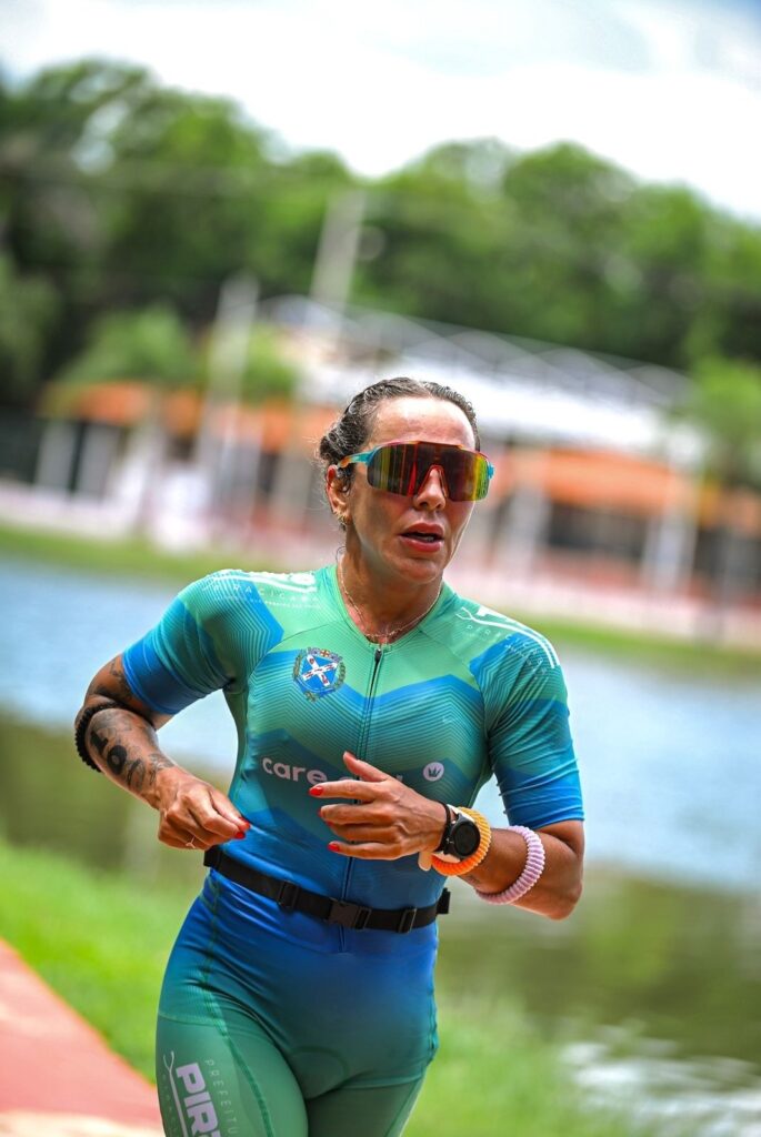 A imagem mostra uma atleta feminina correndo ao ar livre, provavelmente durante uma competição esportiva como um triatlo. Ela está vestindo um uniforme esportivo azul com detalhes em verde e branco, com as palavras "care", "PIRACICABA" e "PREFEITURA" visíveis na parte frontal do traje, indicando que pode representar a cidade de Piracicaba. A atleta usa óculos esportivos grandes e espelhados, tem cabelo preso para trás, unhas pintadas de vermelho e vários braceletes coloridos no pulso. Ela está concentrada e determinada, com expressão focada. O fundo mostra uma paisagem natural com árvores verdes e um corpo d’água sob um céu claro, sugerindo que a corrida ocorre em um parque ou área próxima a um lago. A imagem é nítida e bem iluminada, com o fundo desfocado para destacar a atleta em movimento.