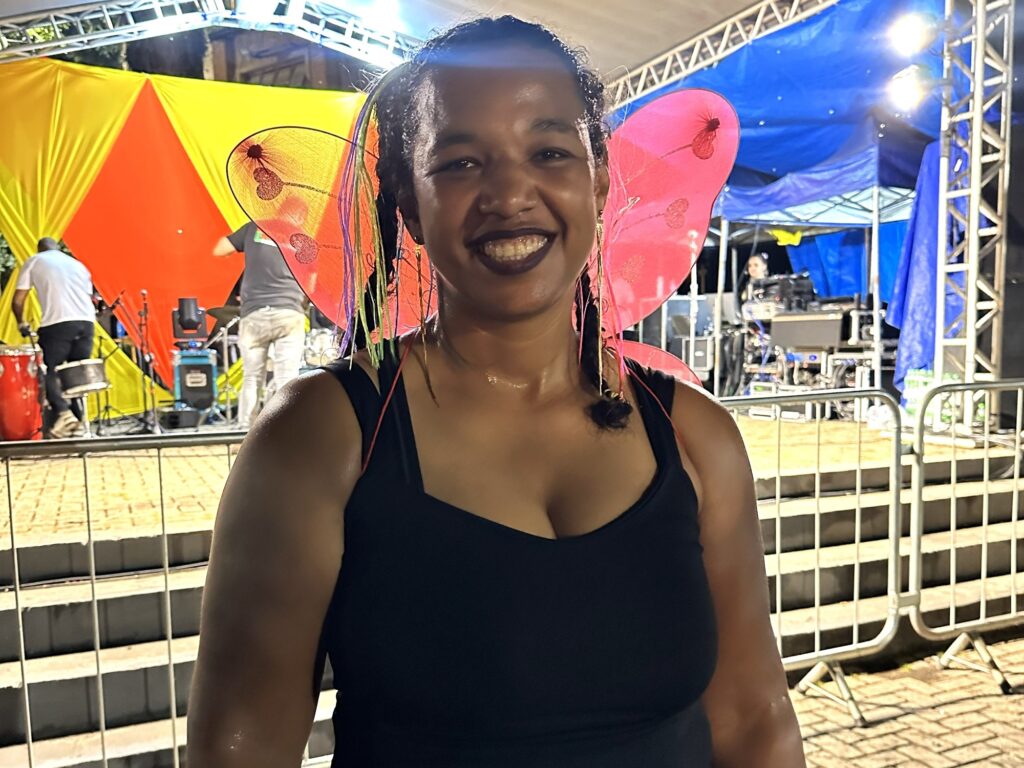 A imagem mostra uma mulher sorridente em primeiro plano, vestindo asas de borboleta cor-de-rosa e uma regata preta. Ela tem cabelo escuro com tranças e detalhes coloridos, e está usando batom escuro. Ela parece estar suada, indicando que pode estar em um ambiente quente ou ativo. Ao fundo, há um palco com equipamentos musicais, como bateria e alto-falantes, além de duas pessoas: um homem tocando bateria e outra pessoa próxima a equipamentos de som. O cenário é noturno e ao ar livre, com iluminação artificial e cores vibrantes (amarelo, vermelho e azul) em tecidos que decoram o local. Há também uma grade metálica separando a mulher do palco. O ambiente sugere um evento festivo, possivelmente um carnaval ou uma festa temática.