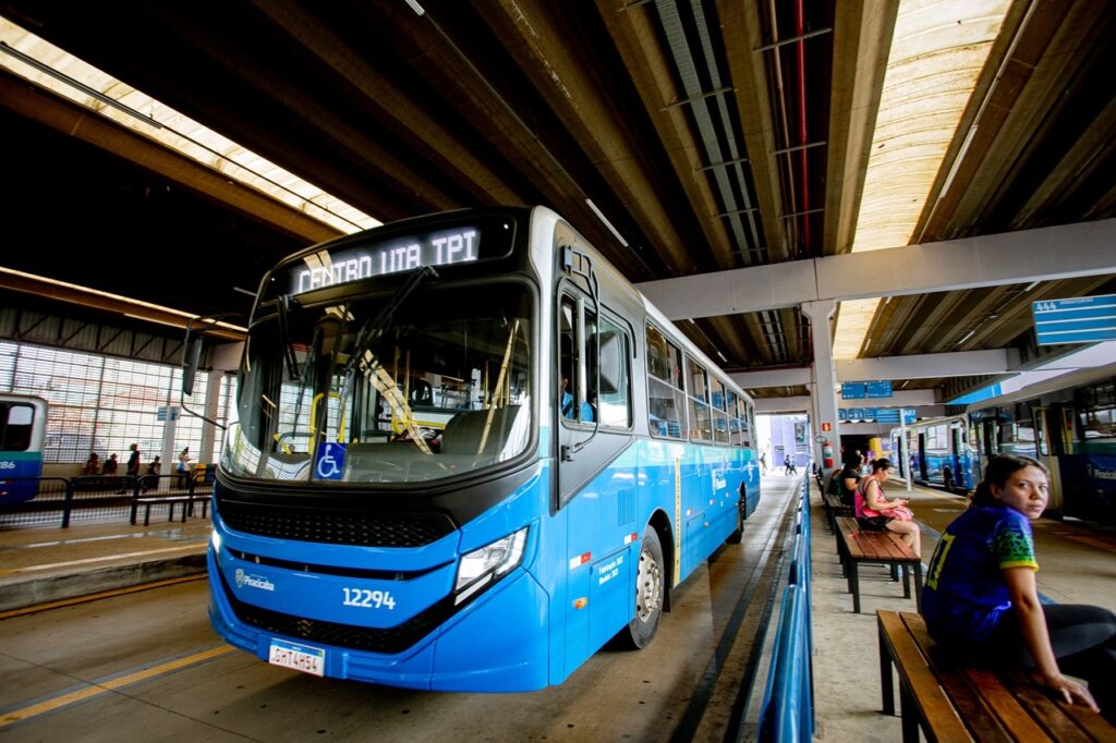 A imagem mostra uma cena em um terminal de ônibus urbano. Em destaque, há um ônibus moderno de cor azul e branca, com o número 12294 visível na parte frontal. No painel eletrônico do ônibus está escrito "CENTRO VIA TPI", indicando seu destino. O terminal possui uma estrutura coberta com vigas metálicas aparentes e iluminação natural que entra por painéis translúcidos no teto. Ao lado direito do ônibus, há vários passageiros sentados em bancos de madeira sob a cobertura do terminal. Entre eles, uma mulher com camisa azul e amarela olha diretamente para a câmera, enquanto outra mulher usa o celular. O ambiente transmite a sensação de um local movimentado, típico de uma estação de transporte público durante o dia. Ao fundo, é possível ver outro ônibus estacionado e algumas placas de sinalização, sugerindo que o terminal atende várias linhas. A imagem é clara e bem iluminada, mostrando detalhes do veículo e do espaço ao redor.