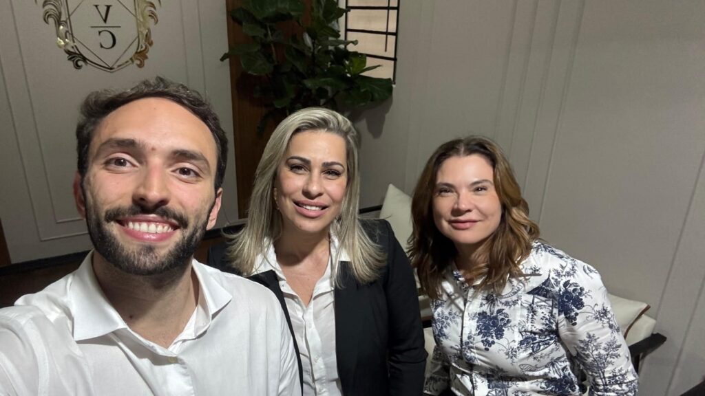 A imagem mostra uma selfie de três pessoas sorrindo, em um ambiente interno que parece ser um escritório ou uma sala de reunião. À esquerda, há um homem com cabelo curto e barba, vestindo uma camisa branca. Ao centro, uma mulher loira usa um blazer preto por cima de uma camisa branca. À direita, outra mulher, de cabelos castanhos, veste uma blusa branca com estampa azul. Ao fundo, há uma planta verde e uma parede com um logo que contém as letras "V" e "C" dentro de um brasão decorativo. O ambiente é bem iluminado, com cores neutras e um clima profissional e acolhedor.