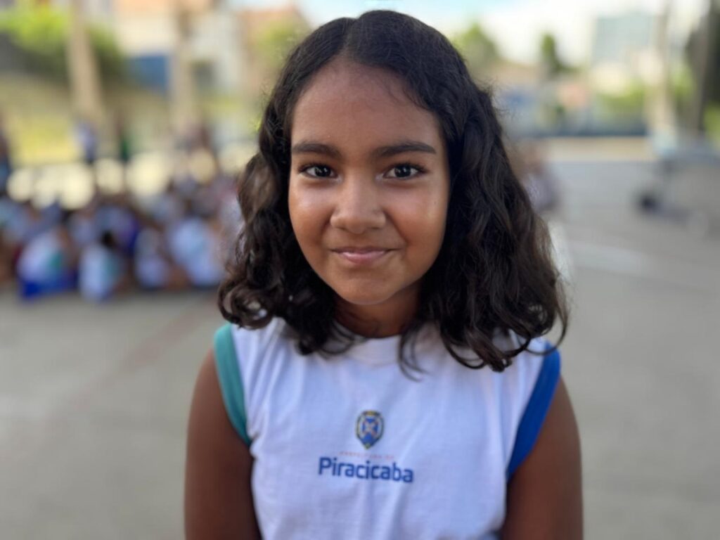 A imagem mostra uma menina jovem, provavelmente estudante, com pele de tom médio e cabelo escuro, ondulado e na altura dos ombros. Ela está olhando diretamente para a câmera com um leve sorriso, transmitindo uma expressão amigável e calma. A menina veste uma camiseta regata branca com detalhes em azul e verde nas bordas das mangas. No centro da camiseta, há um logotipo azul e a palavra "Piracicaba" escrita, indicando que provavelmente ela frequenta uma escola ou instituição localizada nessa cidade. O fundo da imagem está desfocado, com outras crianças sentadas no chão, sugerindo que a foto foi tirada em um ambiente escolar ao ar livre, talvez durante um recreio ou atividade. A luz natural é intensa, iluminando suavemente o rosto da menina e criando um clima alegre e tranquilo na fotografia. A composição destaca a menina em primeiro plano, com o fundo desfocado para dar foco a ela.